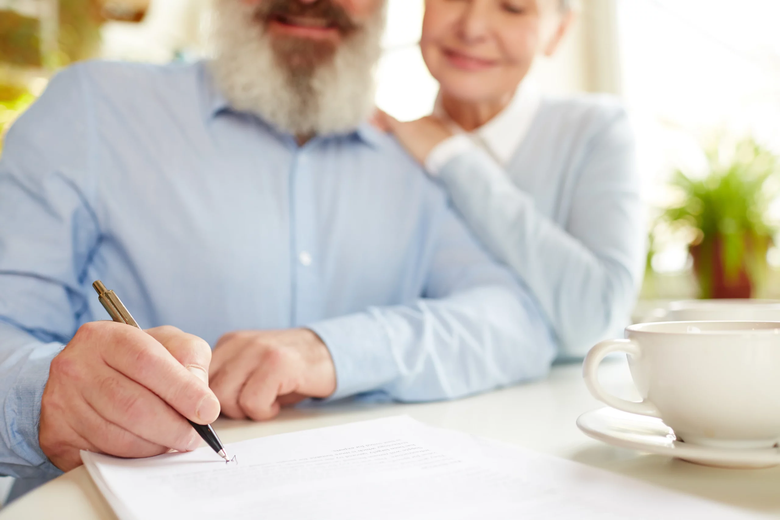 Elderly man signing power of attorney document with partner leaning on his shoulder to support him in warm home-office setting